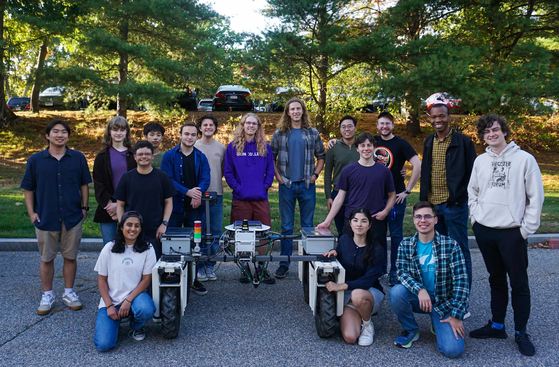 Group of students pose around the PhoenixBot in a gravel parking lot with evergreen trees behind them.