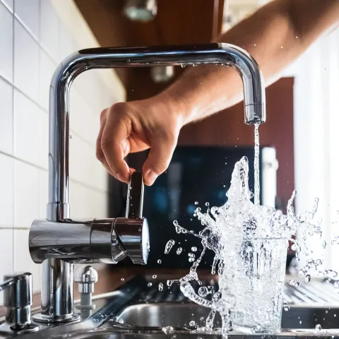 Hand turns on a tap with water splashing out of a glass